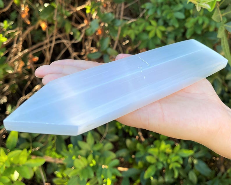 May include: A long, translucent white selenite crystal wand with a smooth, polished surface and a pointed tip. The crystal has a rectangular, faceted shape with a soft, milky appearance that catches the light, showcasing its natural fibrous texture.