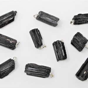 May include: A collection of black tourmaline pendants with silver-colored bails. The raw, natural crystals have a striated texture and varying shapes. The pendants are displayed against a white background, showcasing their dark color and unique forms.