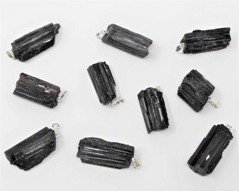 May include: A collection of black tourmaline pendants with silver-colored bails. The raw, natural crystals have a striated texture and varying shapes. The pendants are displayed against a white background, showcasing their dark color and unique forms.