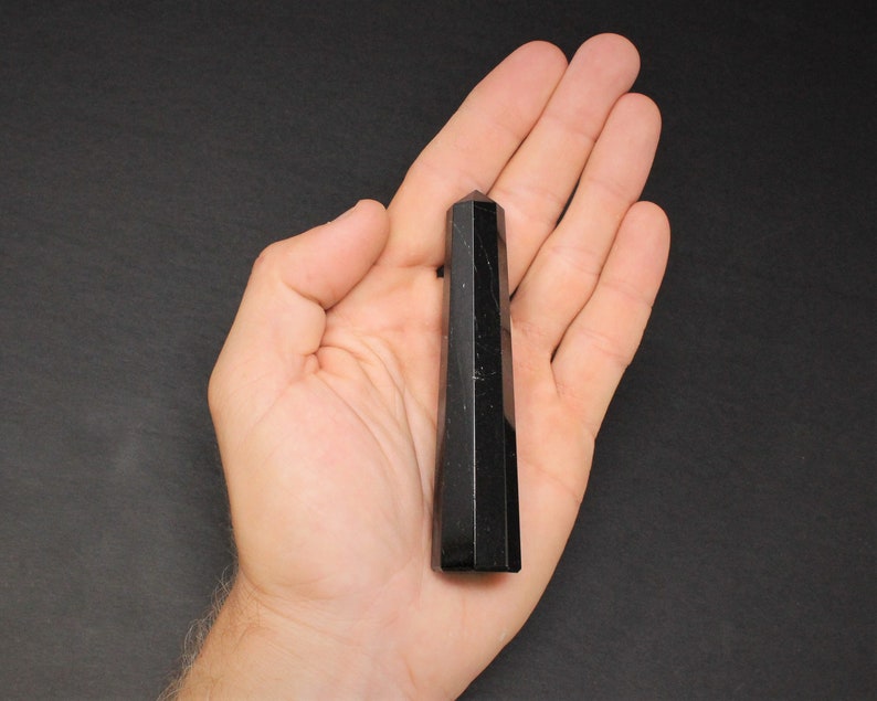 May include: Black obsidian crystal point, a dark, smooth, and pointed stone, held in a hand against a black background.