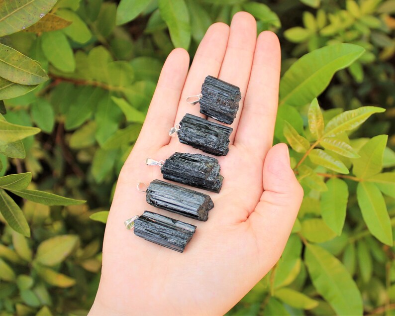 May include: Five raw black tourmaline pendants with silver-colored bails are displayed against a backdrop of green foliage. The tourmaline stones have a rough, natural texture and varying lengths. The pendants are held in a hand, showcasing their dark, opaque color.