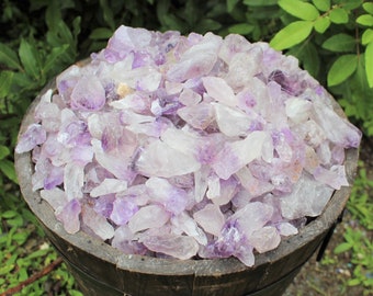 May include: A wooden bowl filled with a large quantity of amethyst crystals. The crystals are a mix of light purple and clear, with some having a more opaque appearance. The crystals are all roughly the same size and shape, and they are arranged in a random pattern.