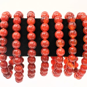 May include: Several strands of red-orange beaded bracelets are displayed on a black velvet surface. Each bracelet is composed of large, round beads. The beads have a glossy finish and a translucent quality, allowing light to pass through. The bracelets are a vibrant accessory.