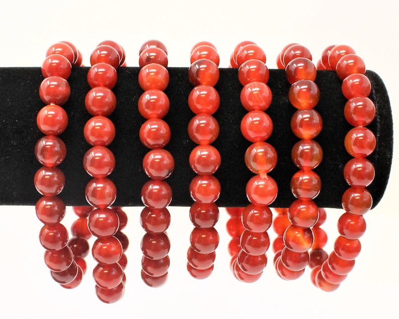 May include: Several strands of red-orange beaded bracelets are displayed on a black velvet surface. Each bracelet is composed of large, round beads. The beads have a glossy finish and a translucent quality, allowing light to pass through. The bracelets are a vibrant accessory.