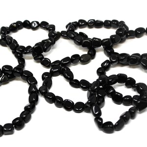 May include: A collection of five circular bracelets made from polished, smooth, black stone beads. Each bracelet is constructed from individual, irregularly shaped, glossy black pebbles strung together to form a continuous, flexible loop.