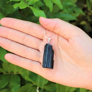 May include: A black tourmaline pendant necklace. The pendant is a rectangular, vertically-oriented crystal with a silver-colored bail and chain. The background is a blurred view of green foliage.