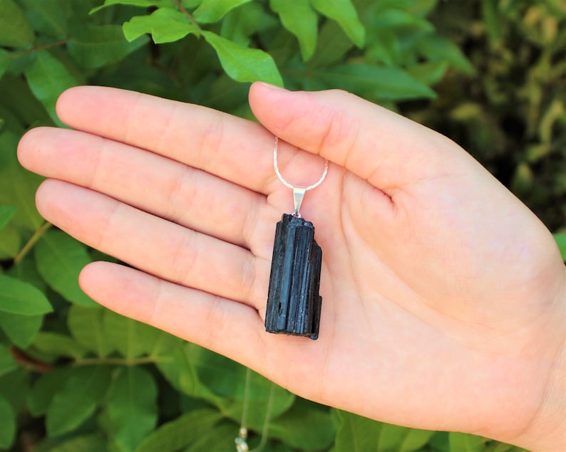 May include: A black tourmaline pendant necklace. The pendant is a rectangular, vertically-oriented crystal with a silver-colored bail and chain. The background is a blurred view of green foliage.
