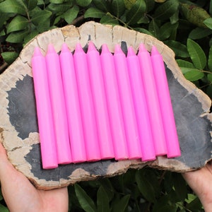 May include: A set of ten pink taper candles arranged on a wooden slab. The candles are all the same size and shape, and they are all pointed at one end.