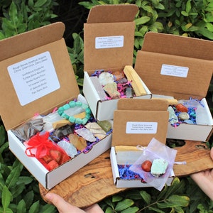 Different sized smallboxes on a wooden board. Each box is full of Crystals, Bracelets, Rough Stones and Tumble Stones. Assorted Mystery Crystals in each Box. Trees in the background.