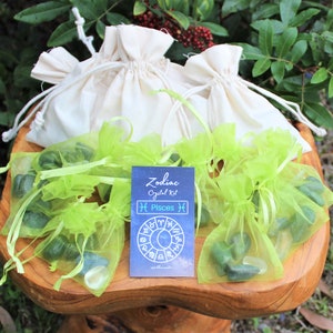 K&ouml;nnte beinhalten: Ein Set gr&uuml;ner Kristallsteine in gr&uuml;nen Organzas&auml;cken, mit einer Karte mit der Aufschrift "Zodiac Crystal Kit Pisces" und einer Grafik eines Tierkreisrades. Die S&auml;cke befinden sich auf einem Holzsockel.