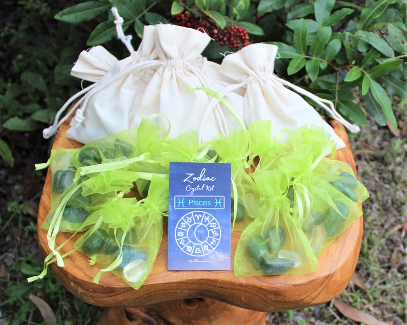 K&ouml;nnte beinhalten: Ein Set gr&uuml;ner Kristallsteine in gr&uuml;nen Organzas&auml;cken, mit einer Karte mit der Aufschrift "Zodiac Crystal Kit Pisces" und einer Grafik eines Tierkreisrades. Die S&auml;cke befinden sich auf einem Holzsockel.