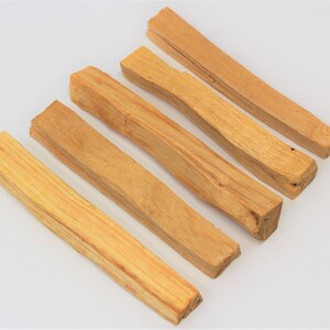 May include: Six light brown Palo Santo wood sticks arranged diagonally on a white surface. The sticks have a natural, rough texture and varying shades of tan and light brown. The ends of the sticks are cut, revealing the wood grain.