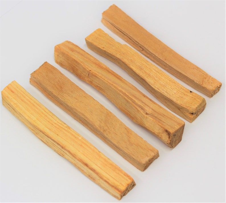 May include: Six light brown Palo Santo wood sticks arranged diagonally on a white surface. The sticks have a natural, rough texture and varying shades of tan and light brown. The ends of the sticks are cut, revealing the wood grain.