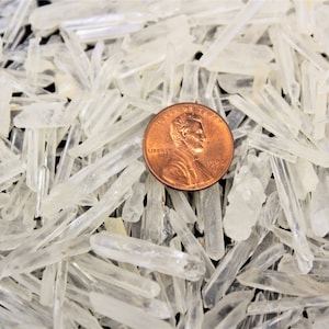 May include: A close-up of a pile of clear quartz crystals, some of which are pointed. A UK penny is included for scale.
