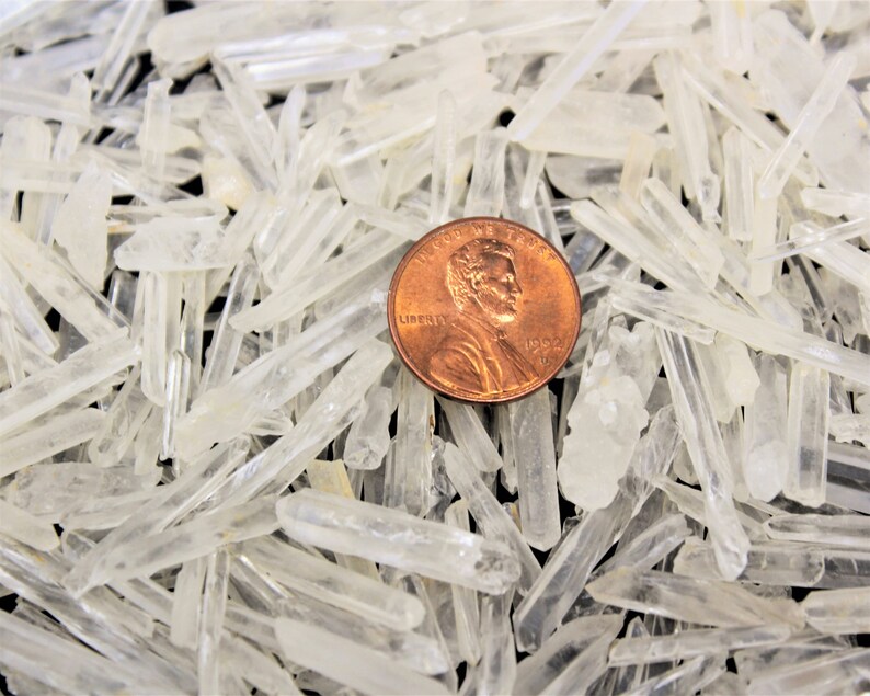 May include: A close-up of a pile of clear quartz crystals, some of which are pointed. A UK penny is included for scale.