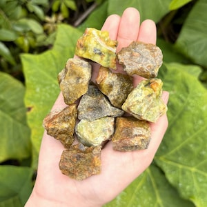 May include: A collection of green and brown rough opal stones in a hand. The stones are various sizes and shapes.