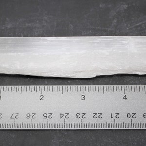 May include: A white selenite crystal stick measuring approximately 4 inches long. The crystal has a rough, natural texture and is displayed next to a ruler for size reference.