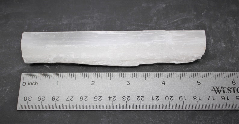 May include: A white selenite crystal stick measuring approximately 4 inches long. The crystal has a rough, natural texture and is displayed next to a ruler for size reference.