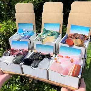 May include: Six small white boxes with lids open, each containing a variety of crystals and gemstones. The boxes are labeled with different crystal themes, including "Positive Energy", "Prosperity", "Strength", "Anxiety & Stress", "Protection", and "Love".