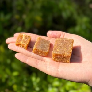 Peut inclure: Trois blocs rectangulaires brun dor&eacute; d'un produit alimentaire sont tenus dans une main. Les blocs ont une surface l&eacute;g&egrave;rement rugueuse et textur&eacute;e et semblent &ecirc;tre solides. L'arri&egrave;re-plan est une vue floue de feuillage vert.
