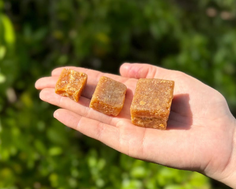 Peut inclure: Trois blocs rectangulaires brun dor&eacute; d'un produit alimentaire sont tenus dans une main. Les blocs ont une surface l&eacute;g&egrave;rement rugueuse et textur&eacute;e et semblent &ecirc;tre solides. L'arri&egrave;re-plan est une vue floue de feuillage vert.