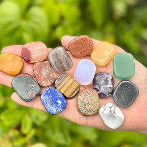 May include: A hand holds a collection of polished gemstones in various colors and patterns. The stones include shades of orange, red, brown, green, blue, and purple. The gemstones are smooth and rounded, with a natural, earthy appearance.