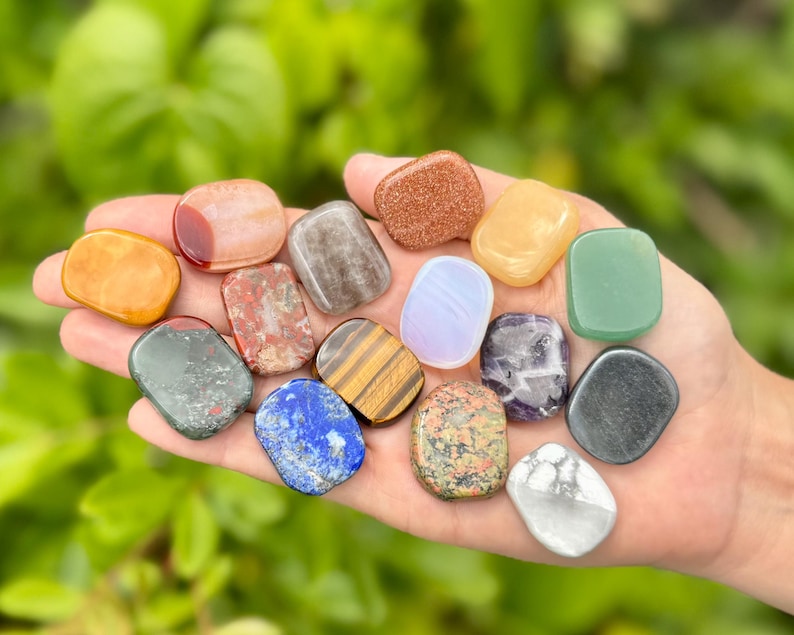May include: A hand holds a collection of polished gemstones in various colors and patterns. The stones include shades of orange, red, brown, green, blue, and purple. The gemstones are smooth and rounded, with a natural, earthy appearance.