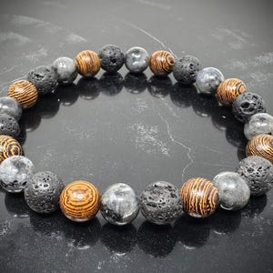 May include: A beaded bracelet featuring a repeating pattern of round, porous black lava beads, smooth grey marbled beads, and brown wooden beads with visible grain patterns arranged in a circular design on a dark reflective surface.