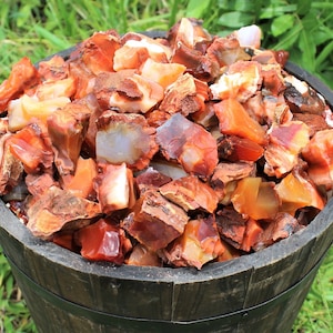 May include: A wooden bucket filled with a large quantity of red and orange carnelian gemstone chips. The chips are various sizes and shapes, with some showing a translucent quality.
