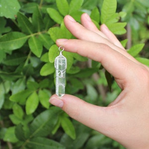 May include: A clear quartz crystal pendant wrapped in silver wire. The pendant is held in a hand against a green leafy background.
