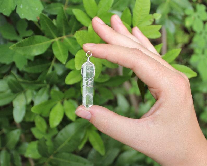 May include: A clear quartz crystal pendant wrapped in silver wire. The pendant is held in a hand against a green leafy background.