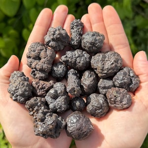 May include: A handful of dark, textured hematite stones. The stones vary in size and shape, with a dark, metallic appearance. The background is a blurred green, suggesting an outdoor setting. The stones are held in open hands.