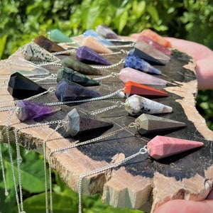 May include: A collection of 18 crystal pendulums with silver chains. The pendulums are made of various crystals, including amethyst, rose quartz, clear quartz, and black obsidian. The pendulums are arranged in a semi-circle on a wooden surface.