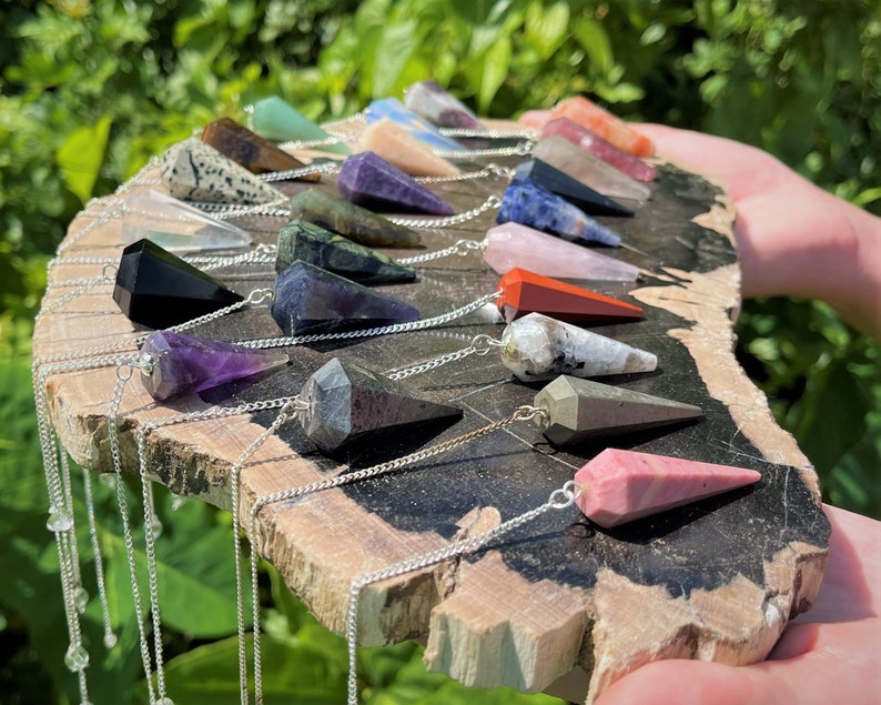 May include: A collection of 18 crystal pendulums with silver chains. The pendulums are made of various crystals, including amethyst, rose quartz, clear quartz, and black obsidian. The pendulums are arranged in a semi-circle on a wooden surface.