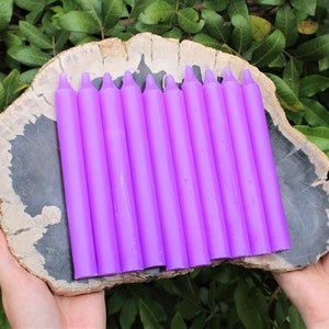 May include: Ten purple taper candles arranged on a wooden slab. The candles are approximately 8 inches long.