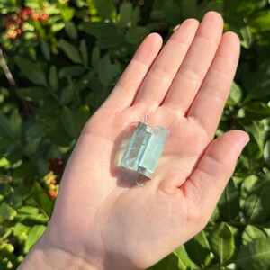May include: A rectangular, light blue aquamarine pendant with silver-colored metal accents. The gemstone is held in a hand against a backdrop of green foliage. The pendant is approximately 1.5 inches long.