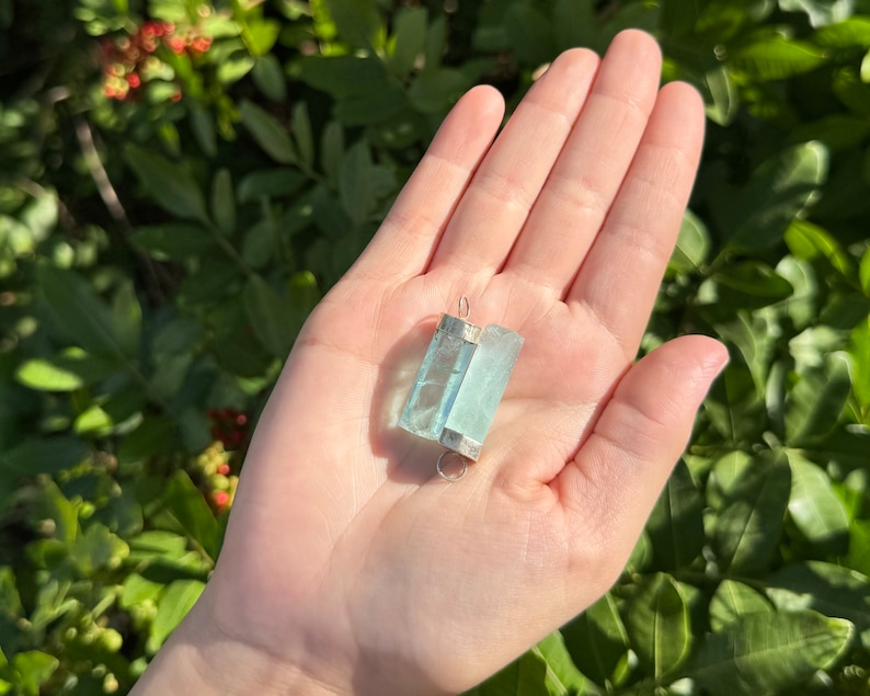 May include: A rectangular, light blue aquamarine pendant with silver-colored metal accents. The gemstone is held in a hand against a backdrop of green foliage. The pendant is approximately 1.5 inches long.