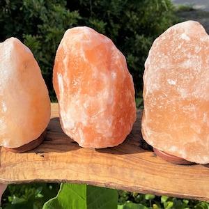 May include: Three Himalayan salt lamps on a wooden base. The lamps are different sizes and shapes, and they are all a light orange color.