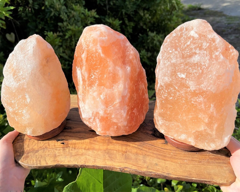 May include: Three Himalayan salt lamps on a wooden base. The lamps are different sizes and shapes, and they are all a light orange color.