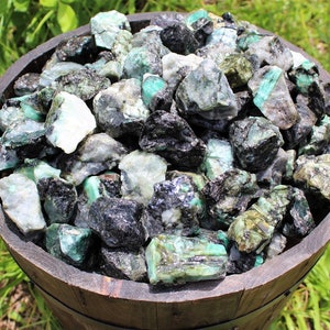 May include: A wooden bowl filled with rough, green emerald crystals. The crystals are a mix of dark and light green, with some showing black inclusions.
