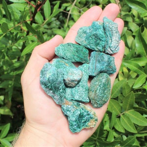 May include: A collection of rough, green, and blue-green malachite stones in various sizes and shapes. The stones are held in a person's hand.