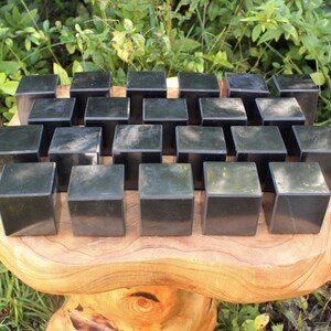 May include: A collection of 25 black obsidian cubes arranged in a grid pattern on a wooden surface. The cubes are smooth and polished, with a slight sheen.