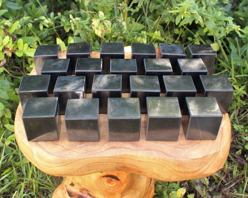May include: A collection of 25 black obsidian cubes arranged in a grid pattern on a wooden surface. The cubes are smooth and polished, with a slight sheen.