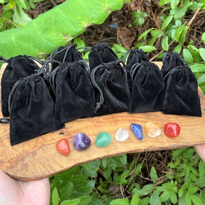 May include: Seven small, smooth, polished stones in various colors, including red, orange, yellow, green, blue, indigo, and violet, are arranged in a row on a wooden surface. The stones are in individual black velvet pouches.