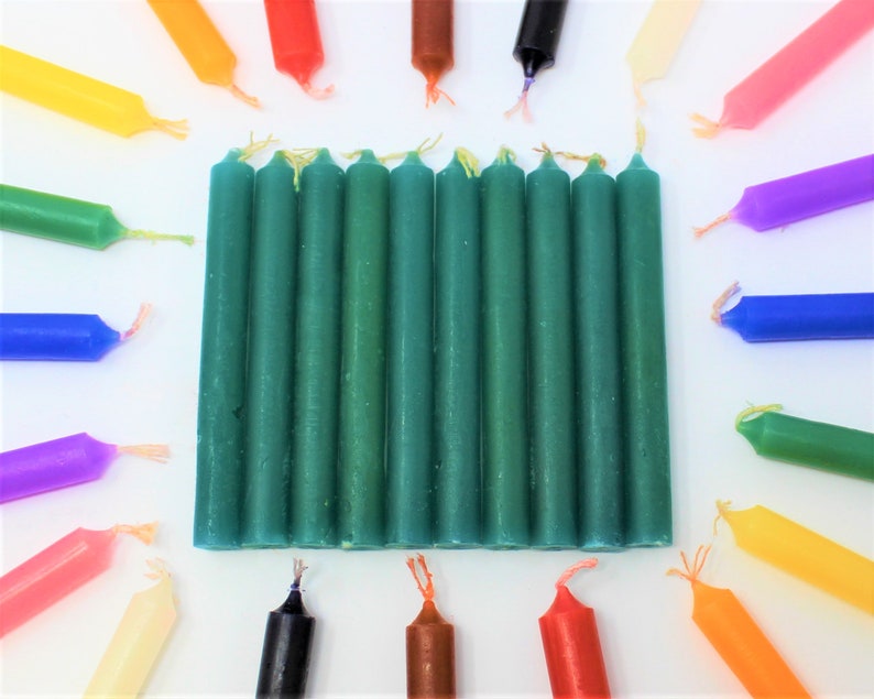 May include: A group of 15 green pillar candles arranged in a rectangle on a white surface. The candles are surrounded by a variety of colored candles, including red, orange, yellow, green, blue, purple, and brown.