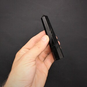 May include: A black obsidian crystal point, also known as a wand, is held in a hand against a black background.