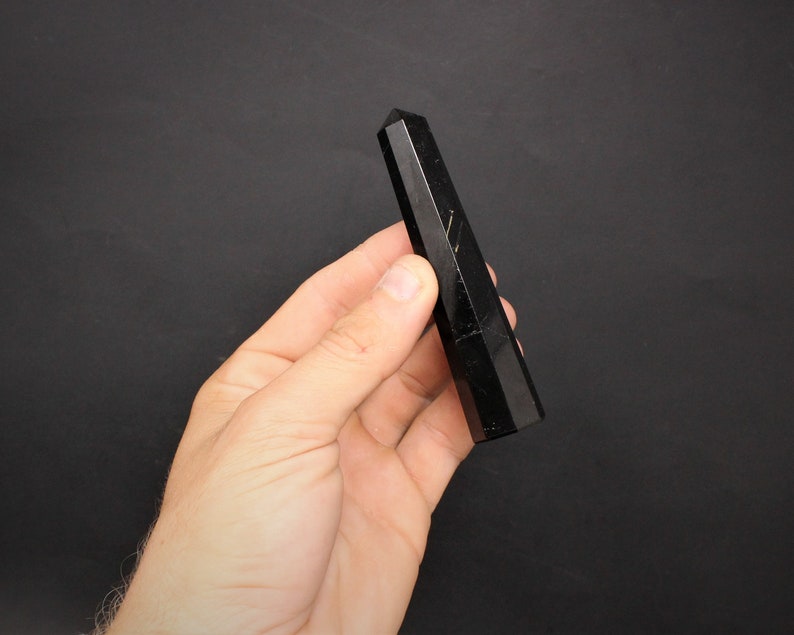 May include: A black obsidian crystal point, also known as a wand, is held in a hand against a black background.