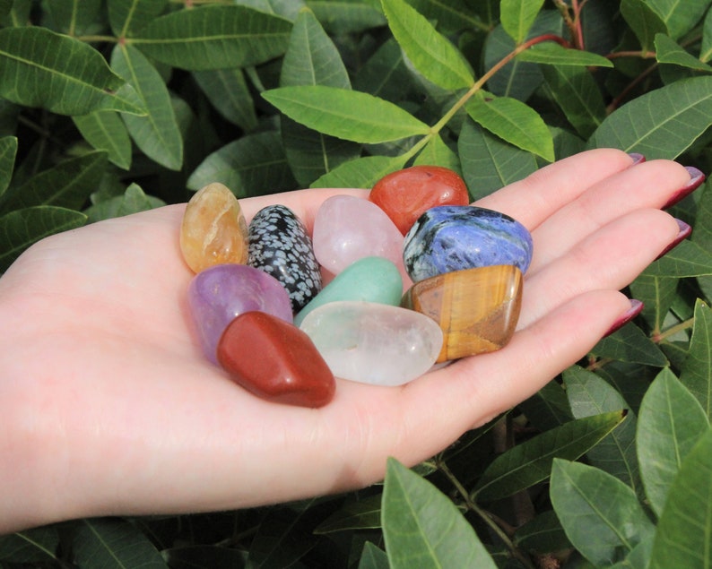 May include: A collection of tumbled stones in various colors, including red, orange, yellow, green, blue, purple, and white. The stones are smooth and polished, and they are arranged in a hand.