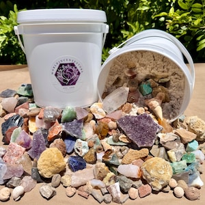 May include: Two white plastic buckets filled with a variety of raw, unpolished gemstones and minerals in shades of purple, blue, green, orange, and tan. One bucket features a purple logo with the text WORLDINCENSESTORE EST. 2006.