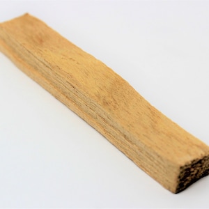 May include: A rectangular piece of light brown palo santo wood. The wood has a rough, textured surface and a natural, earthy appearance. The end of the wood is dark brown.